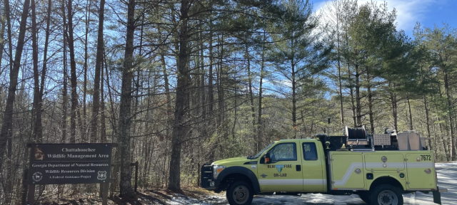 A yellow BLM fire truck parked on a road next to woods on a sunny day. Sign reads Chattahoochee Wildlife Management Area. 