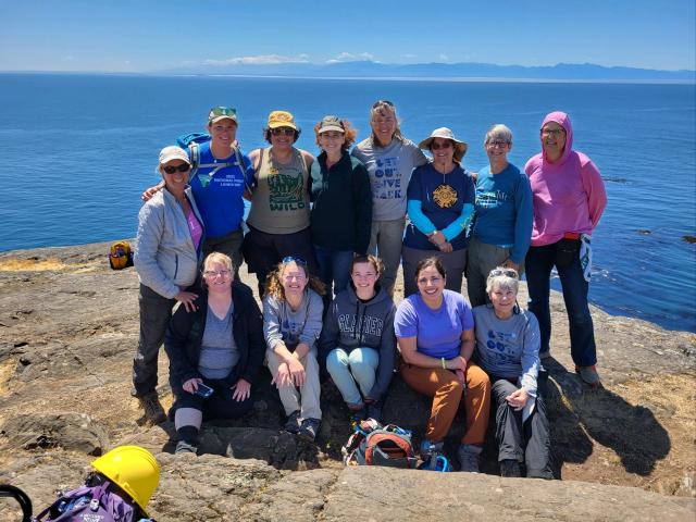 A group of 13 people smile, some sitting and some standing, on some rocks on the edge of blue water. 