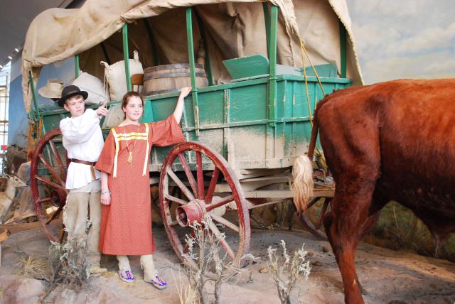 two children in old fashioned garb standing next to display of a covered wagon