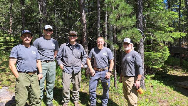 Five caucasian men in blue shirts and green pants pose for the camera