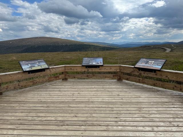 A photo showing a wooden platform with interpretive signs sitting on wooden rails around the platform, which is overlooking rolling hills, a blue sky with white clouds, and mountains in the distance.