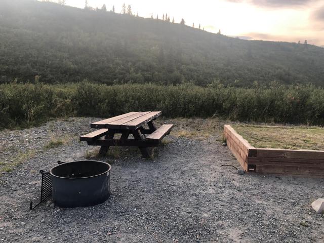 A photo showing a grill, picnic table, and elevated tent pad on a packed gravel campsite with a spruce-covered hill in the background.