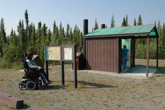 A photo showing an accessible restroom, covered by a green shade structure. A person in a wheelchair looks at a sign with a map of the Paxson Lake Campground. Behind the restroom is a stand of dark green spruce trees.