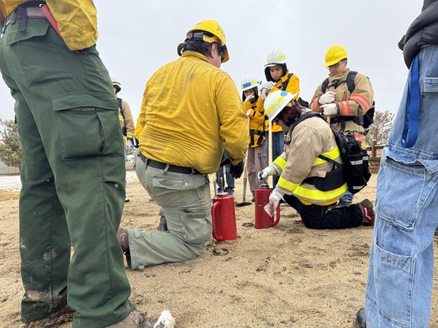 An adult man and a teenager kneel on the ground and examine red fire torches while other teenage people watch. Everyone is wearing firefighter gear.