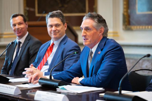 Interior Secretary Doug Burgum sits at a table with microphones. Two men, also dressed in suits, sit to his right.men