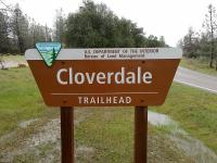 Cloverdale Trailhead sign with paved road, green grass, and trees in the background.