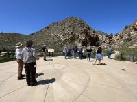 A group of people visit an interpretive site with a hill in the background and blue skies.