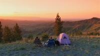 Two people and two dogs sit next to a tent looking out over a valley at sunset. 