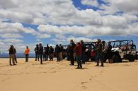 a crowd of people stand in front of off-highway vehicles on a sand dune