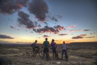 Four mountain bikers stop alongside a trail with their bikes to admire a colorful sunset lighting scattered clouds in pink hues.