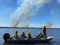 A boat carries BLM and USFWS personnel during the Lathrop Bayou prescribed burn. Heavy smoke can be seen rising against the blue sky from the trees across the water in the distance.