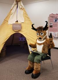Seymour antelope mascot sits with a book in front of a replica tipi. 