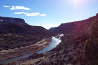 A wide photo of a river running through a deep brown canyon, with blue skies and bright sun in the background.