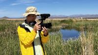 The image shows a man wearing a brimmed hat and yellow jacket standing near a wetland area with mountains in the background. He is looking down into the viewfinder of his video camera with headphones around his neck. 