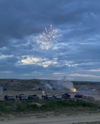 Several vehicles are parked in an open, gravel pit area while a firework goes off above.