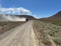 A front loader tractor creates a dust cloud over an unpaved road with mountains in the background.