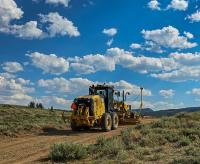 A large, yellow piece of road equipment moves along a dirt road under a blue sky filled with fluffy clouds.