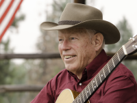 A man with a cowboy hat wearing a red shirt holds a guitar. 