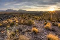 The sun sets over a rocky landscape and scattered desert brush.