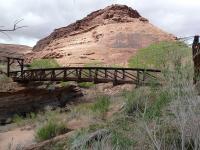 he photo shows a dark brown metal bridge spanning a small canyon with a large sandstone rock formation in the background.