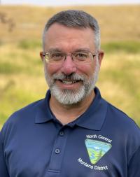 Head-and-shoulders photo of a man with dark-brown and salt-and-pepper silver hair and full beard, fair-complexioned, wire-rimmed glasses, navy-blue shirt with BLM logo, grassy hillside in the background.
