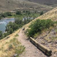 A dirt trail curves around the side of a hill with a river and trees beyond.