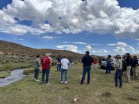 A group of people stand near a creek in green grass under blue sky and clouds