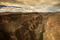 A massive gorge with a river at the bottom cut through a desert landscape, with clouds in the background.