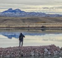 A person fishes from a rock jetty surrounded by the water of a scenic reservoir with partially snow-covered mountains for the backdrop.