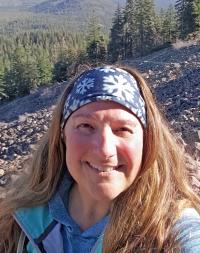 Photo. Foreground: head-and-shoulders view of a woman, smiling, with reddish-brown, long hair, fair-skinned, wearing a snowflake-printed headband and blue zip-up jacket. Background: outdoors, daytime, sunny, forest and rocky hillside.