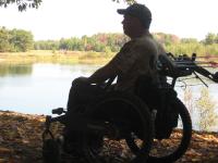 A photo showing a man in a wheelchair next to a river. He’s wearing camouflage and has an adaptive bow mount on his wheelchair.