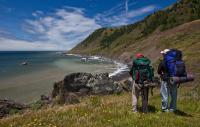 Two people wearing hiking backpacks look out at the beach below a hill.
