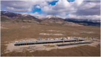A landscape scene with brown hills and a partly cloudy sky. The foreground is taken up by a long black low-slung structure, a geothermal power plant.