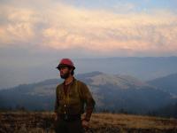 a firefighter wearing a hard hat and Nomex poses in front of a smokey landscape of hills