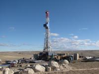 Oil drilling rig set against a clear blue sky with scattered clouds. The foreground features large rocks and vehicles.