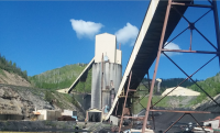 Industrial mining facility with tall silos and conveyor belts set against green hills under a clear blue sky.