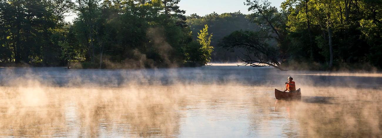 Fisherman wearing orange life jacket and tan hat at sunset with a blanket of fog surrounding the boat.