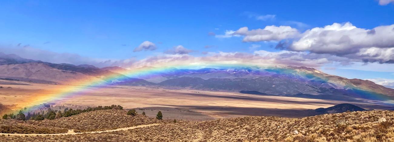A rainbow curves above a landscape of brush and plains, with mountains in the distance.