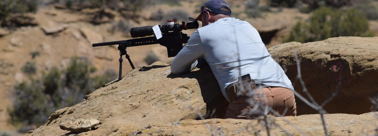 A photo of a man leaning on a rock in the desert, aiming a firearm into the distance.
