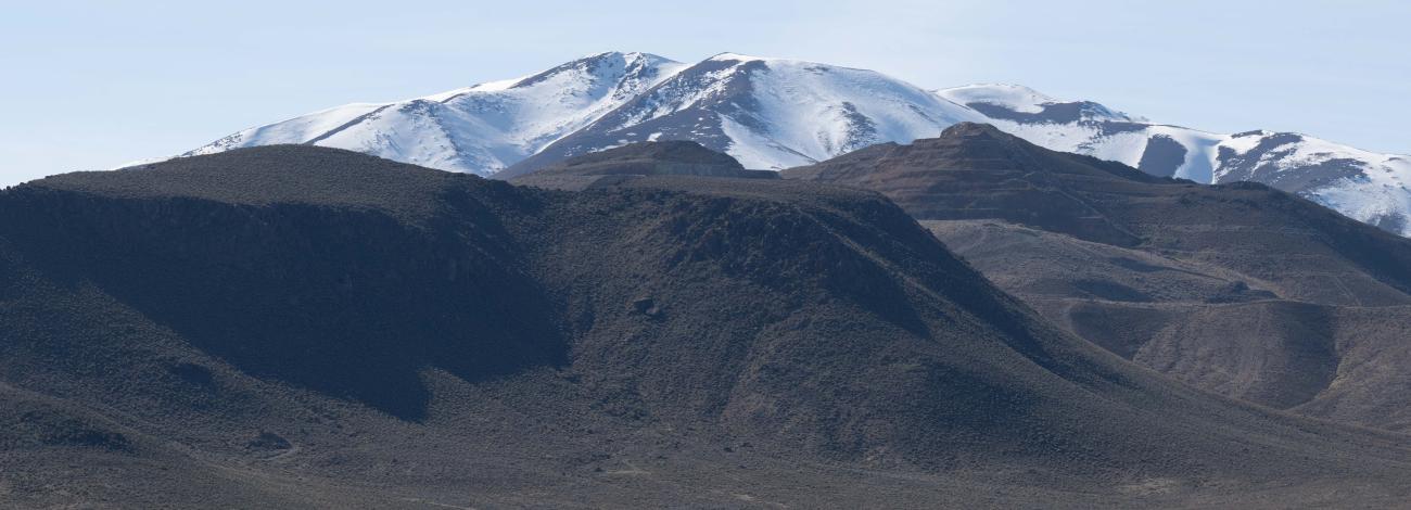 In the foreground a truck leaves a dust trail as it passes from left to right beneath a snowcapped mountain near Battle Mountain, Nevada March 20, 2024.