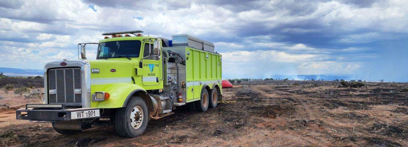 A light green wildland fire truck or water tender is parked on desert scorched black from a wildfire.