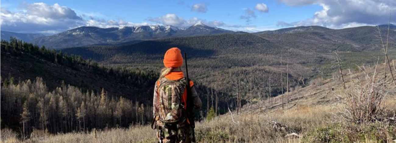 a photo of a child wearing hunter orange and camouflage standing on a hill with sprawling mountain views under a blue sky with puffy white clouds.