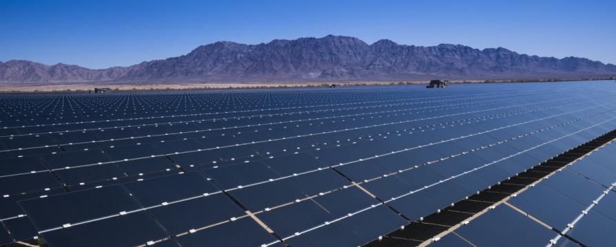 Image shows numerous rows of solar panels in the middle of a desert field surrounded by mountains under a clear sky.