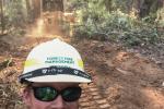 A person wearing sunglasses and hardhat takes a selfie in a forest with an excavator digging fireline behind him.