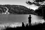 The black and white photo shows an adult standing next to two sitting children. They are on the shore of a small lake with a tree-covered hill in the background.