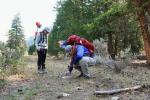 Two BLM ID archaeologists, one standing and one squatting, are surrounded by trees and are documenting surface artifacts along the Salmon River.