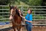 A woman smiles at the camera as she stands next to her brown horse with white facial markings inside of an outdoor pen