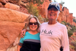 A couple stand on a trail in front of red rocks. 