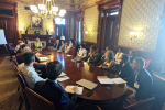 Group of people sit around a table looking at Dr. Jane Lubchenco, located in a large room.