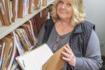 A woman stands next to a file cabinet packed with file folders, holding a file folder open.